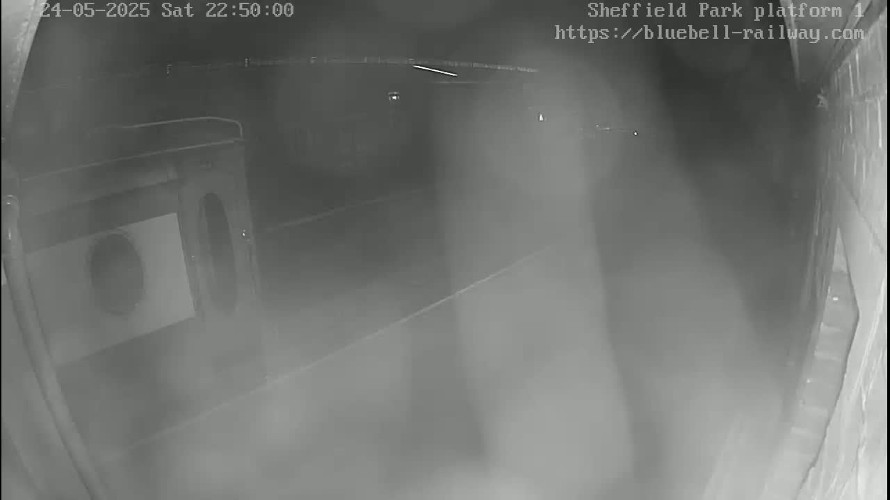 A nighttime low-light security camera view shows a dimly lit, empty platform area with a structure resembling a large machine or cabinet to the left.