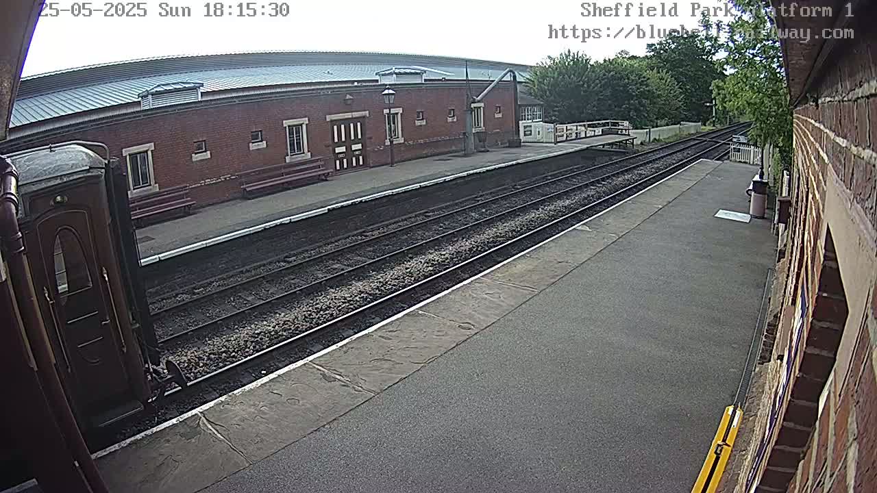 A train car is parked at a railway station platform on a partly cloudy day, with train tracks extending into the distance past a brick building.