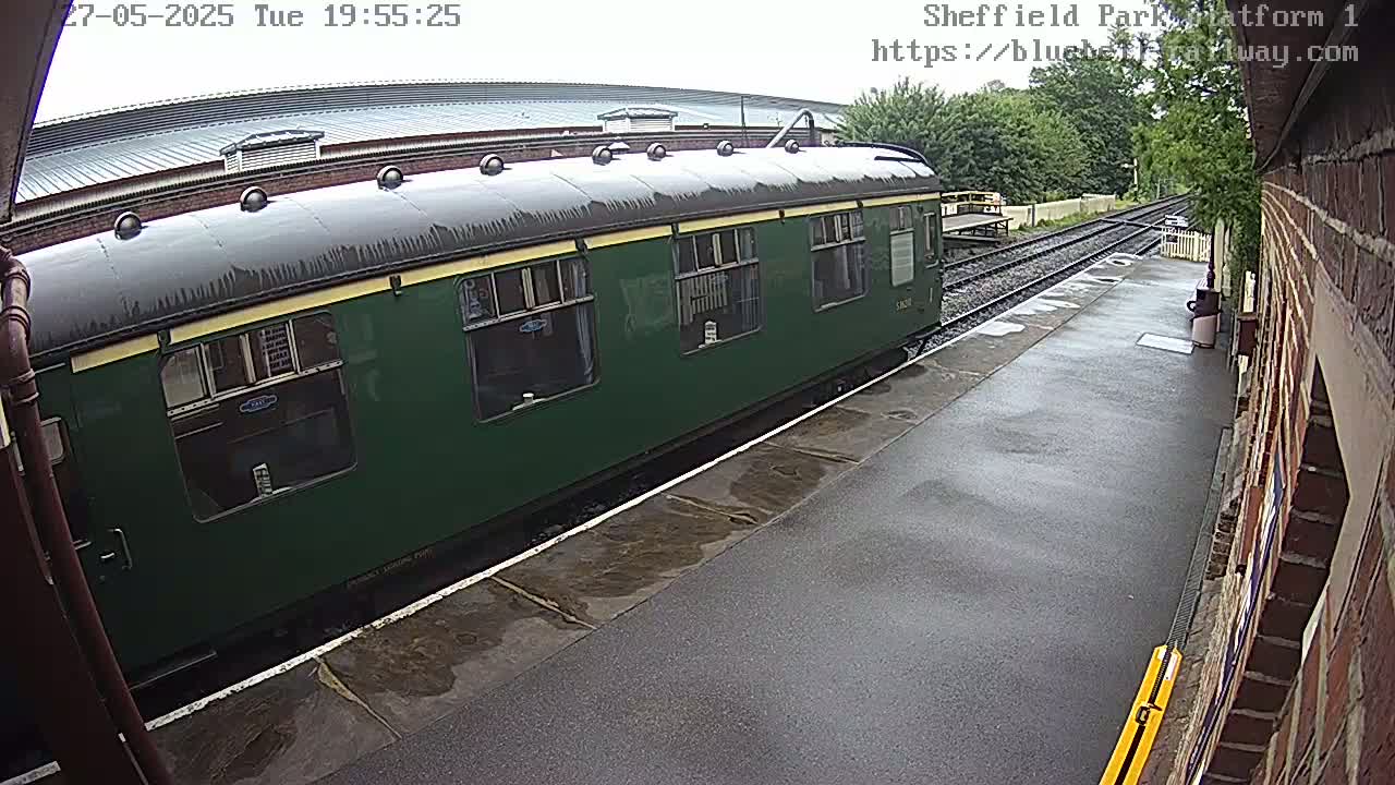 A dark green passenger train car sits on a wet train platform next to a brick building.