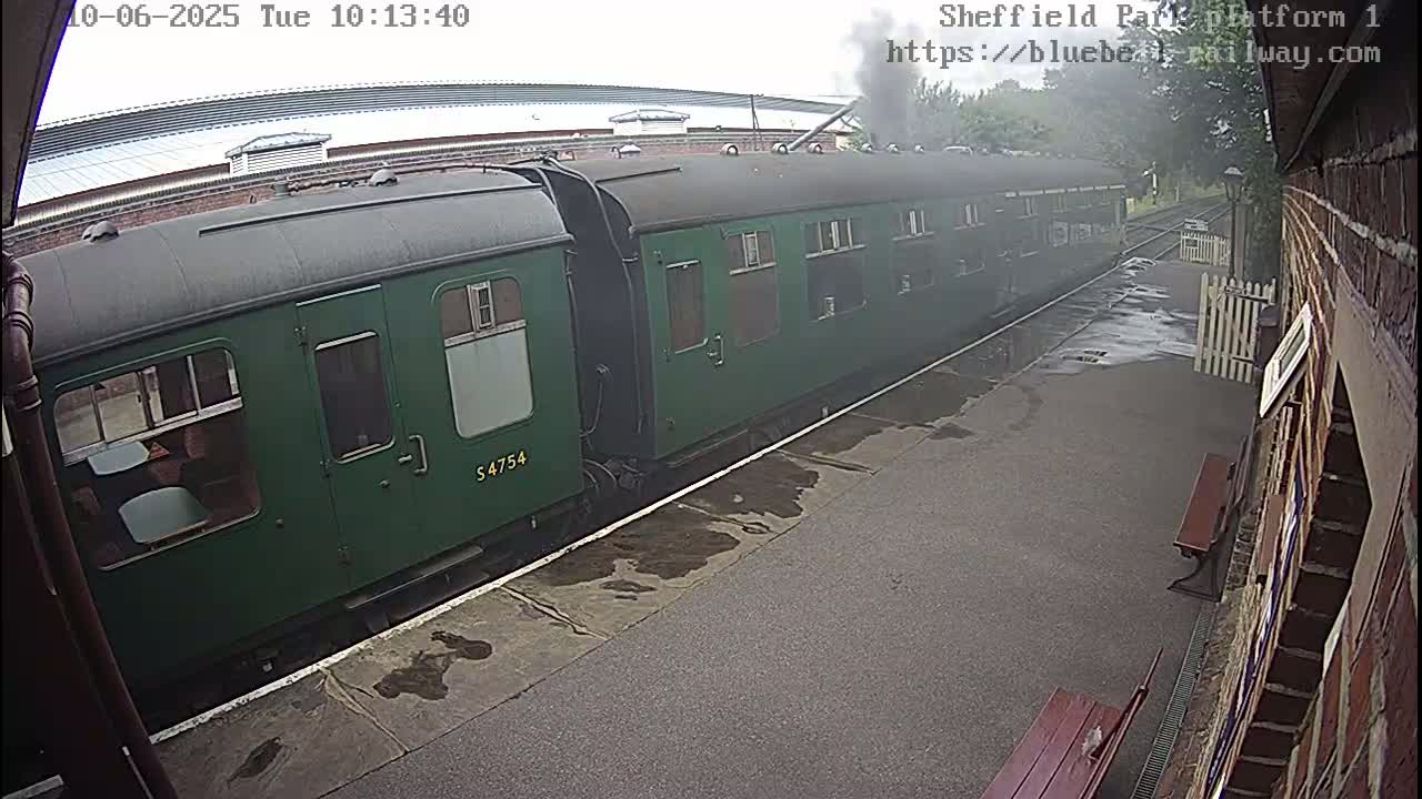 A long, dark-green passenger train sits at a wet train platform on an overcast day.