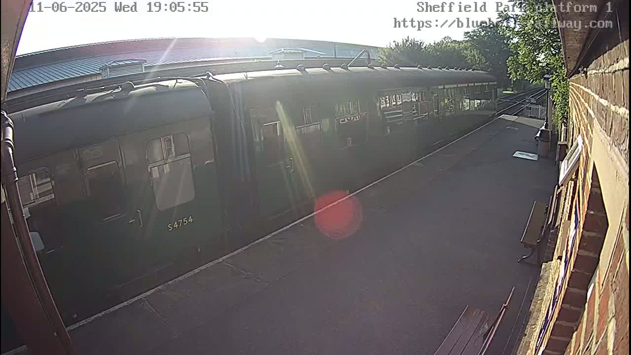 A dark-green passenger train is at a station platform in bright, sunny weather.