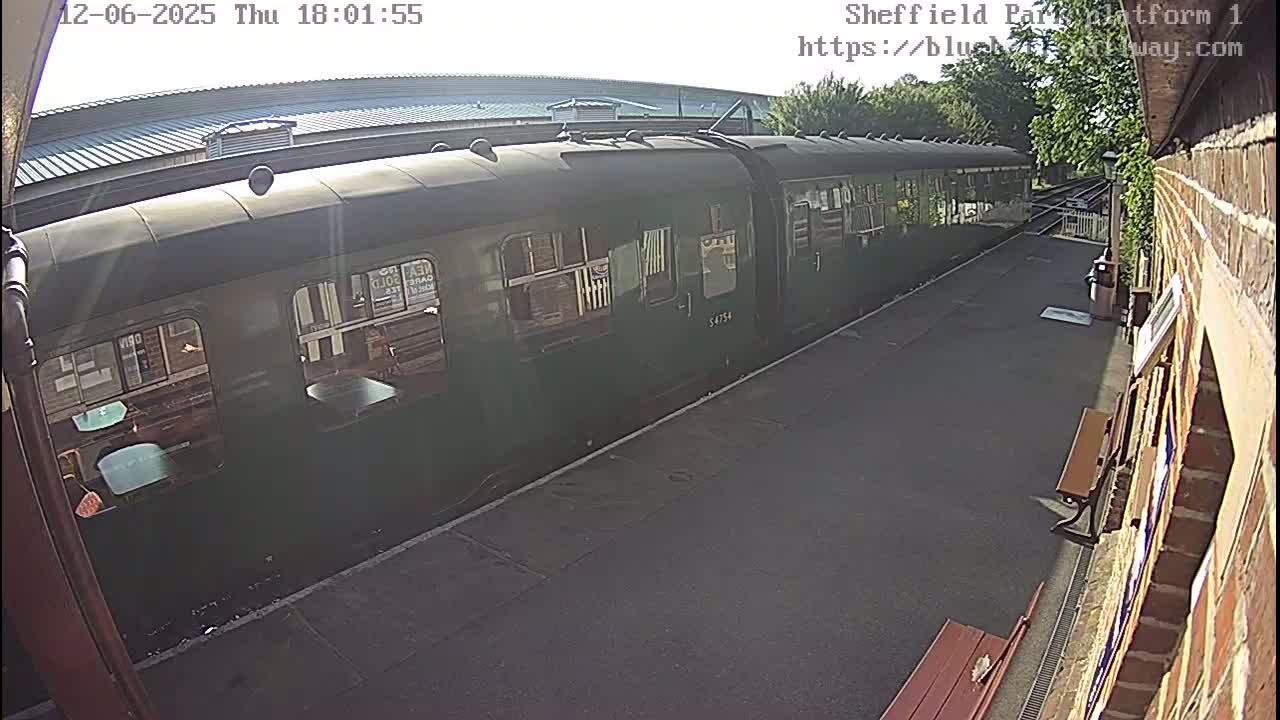 A long, dark-green passenger train sits at a station platform on a sunny day.