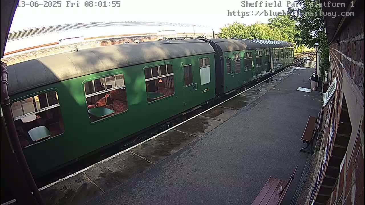 A long, dark-green passenger train sits at a station platform on a partly sunny day.