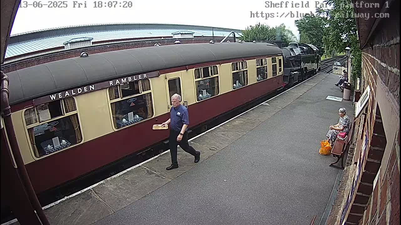 A red and cream passenger train is at a station platform on an overcast day, with a man walking away from it and a woman sitting on a bench.