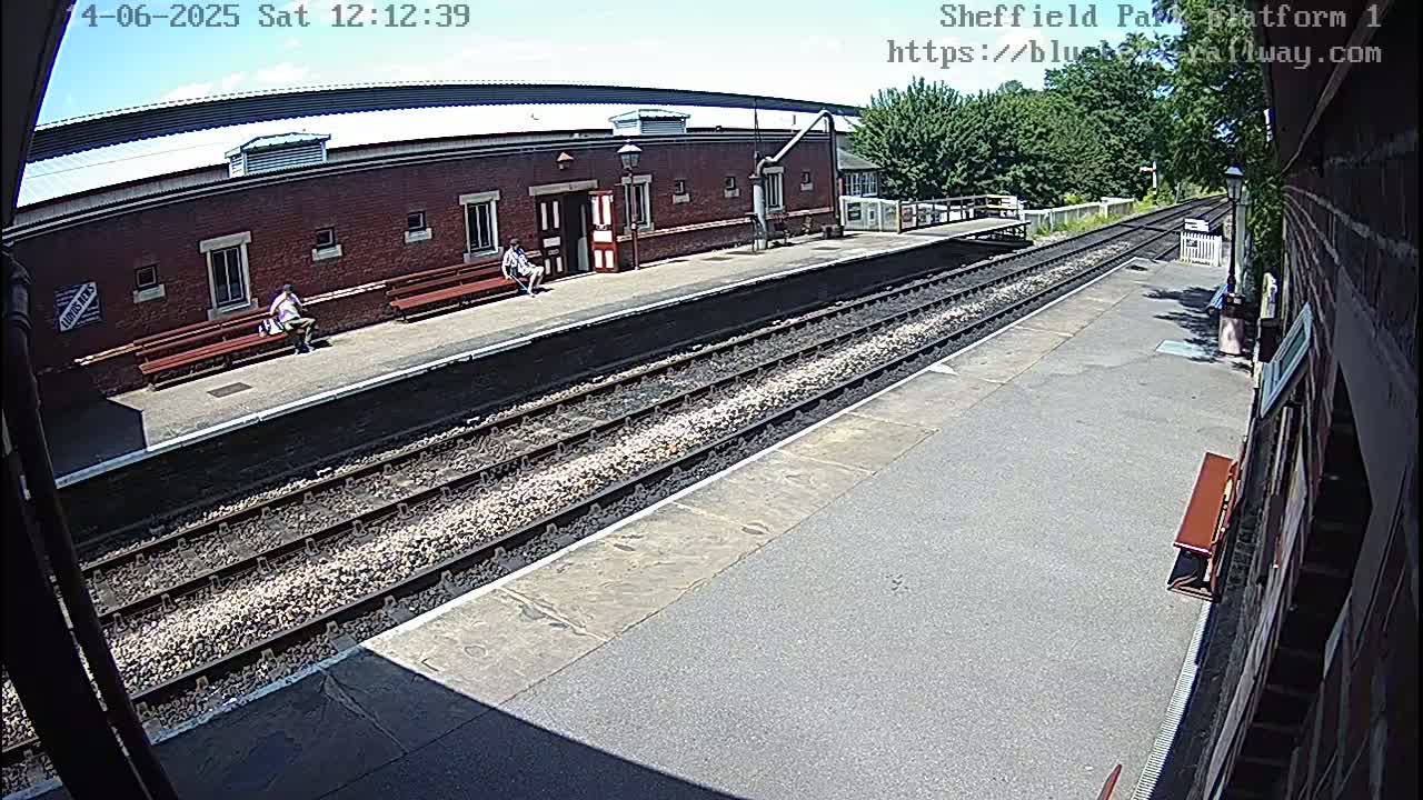 A sunny view of a train platform with two people sitting on benches, multiple train tracks, and a brick building in the background.