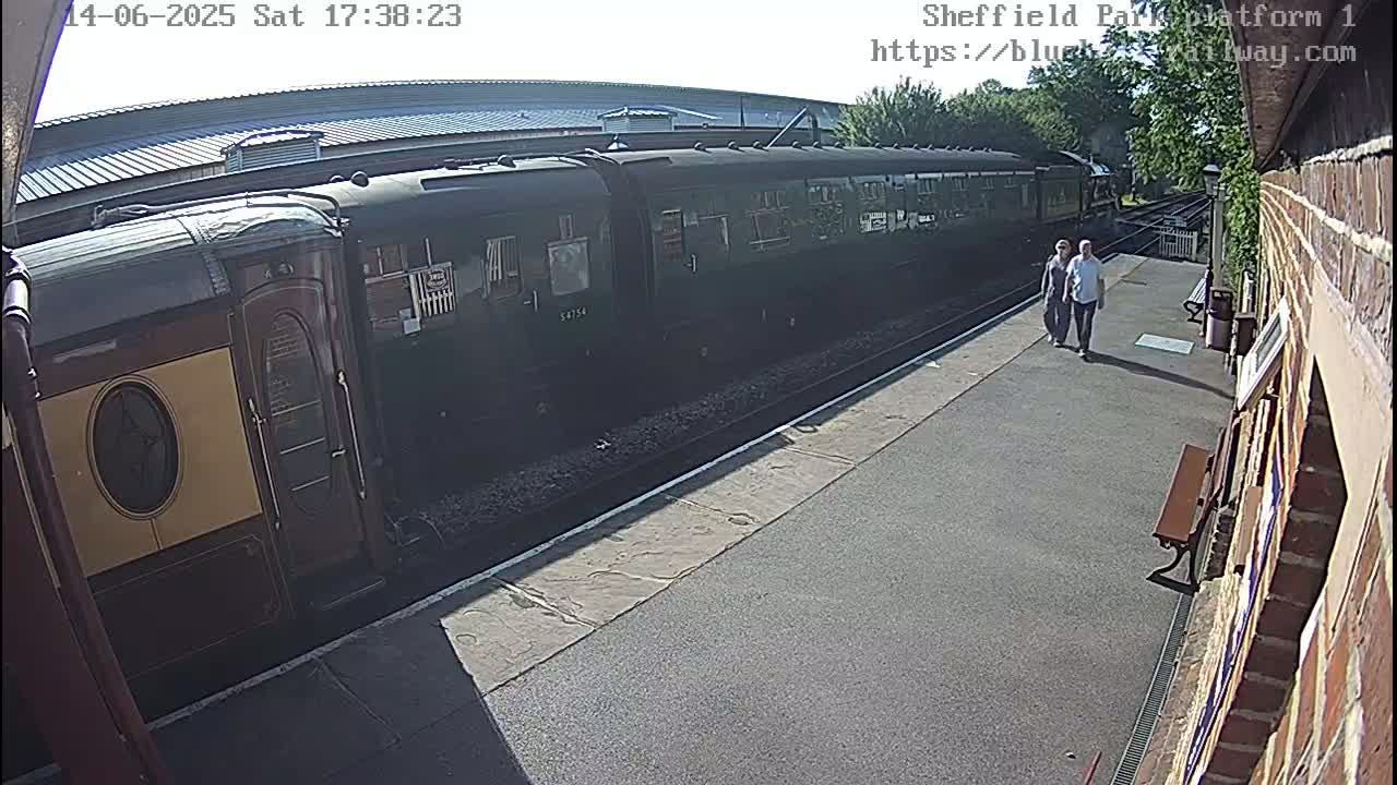 A long, dark-green passenger train is at a station platform on a sunny day, and two people are walking away from it.
