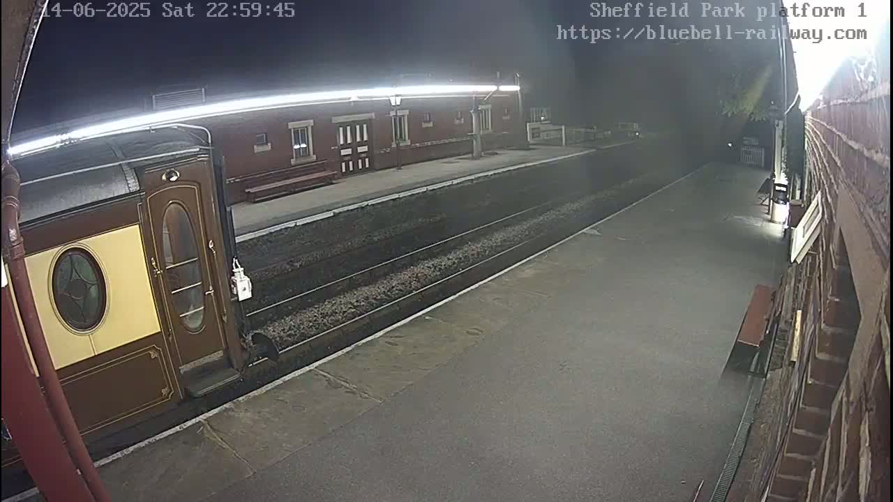 A long, dark-green passenger train is at a station platform on a sunny day, and two people are walking away from it.