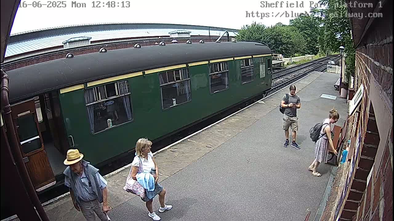 A dark-green passenger train is stopped at a station platform on a partly sunny day, where several people are standing near it and a brick building.