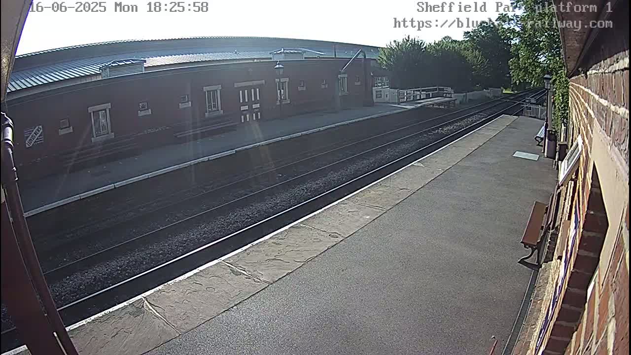 A mostly sunny view of a train platform with a brick building and train tracks.