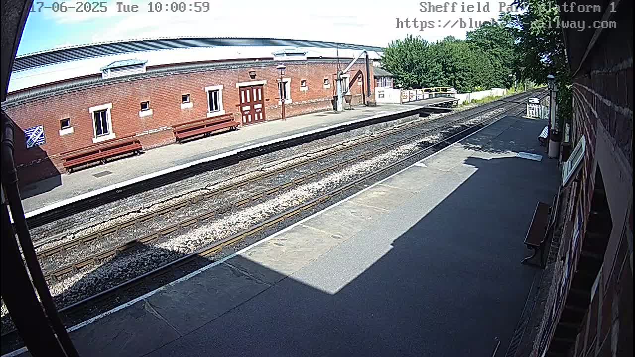 A sunny view of a train platform with a brick building, benches, and train tracks.
