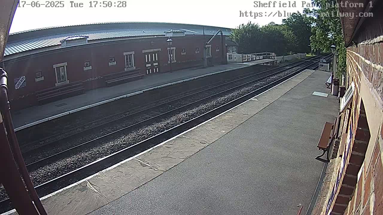 A train platform on a sunny day shows a red brick building, train tracks, and a partially visible bench.