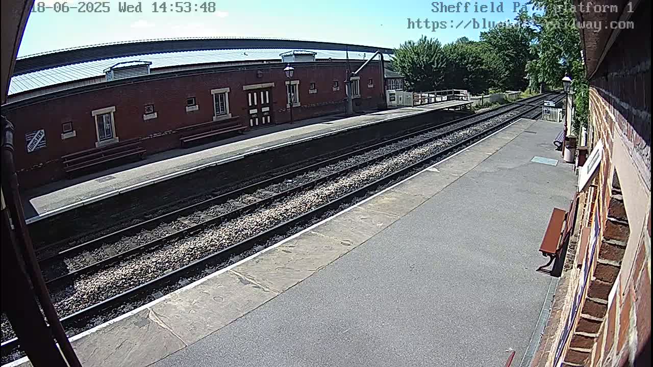 A sunny view of a train platform with two sets of train tracks and a brick building in the background.