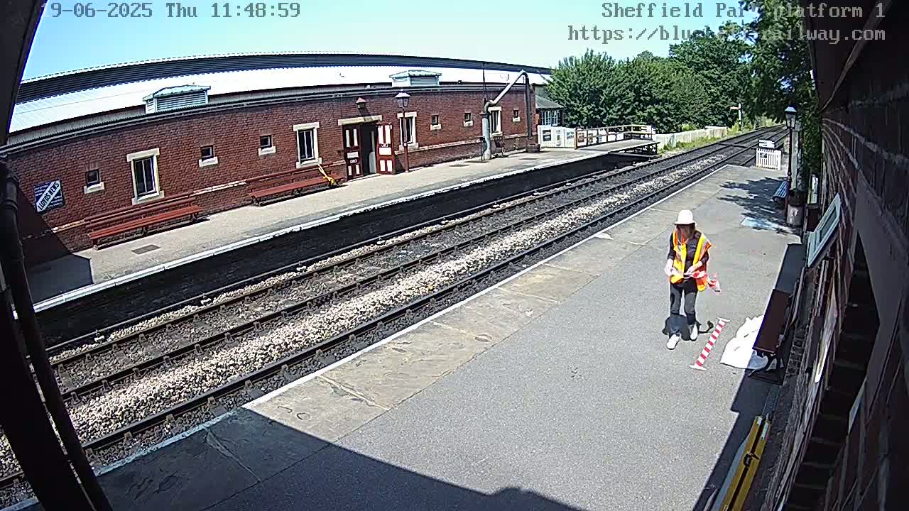 A person in a bright orange safety vest stands on a sunny train platform next to railway tracks and a brick building.
