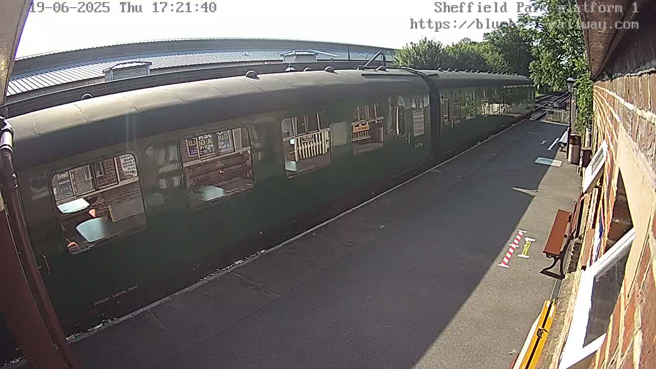 A long, dark-green passenger train sits at a station platform on a sunny day.