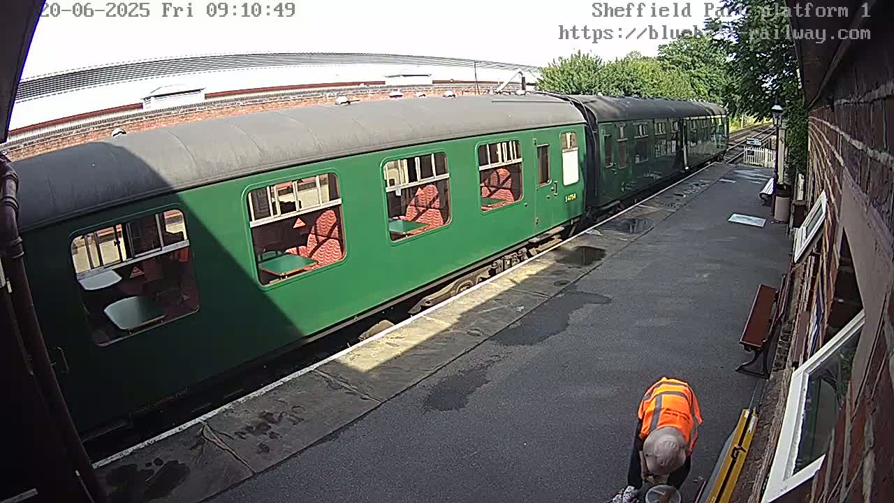 A green passenger train car is parked at a station platform on a partly sunny day, and a person in an orange safety vest is seen cleaning near the train.