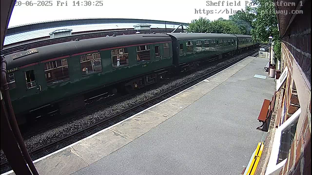 A long, dark-green passenger train sits at a station on a sunny day.