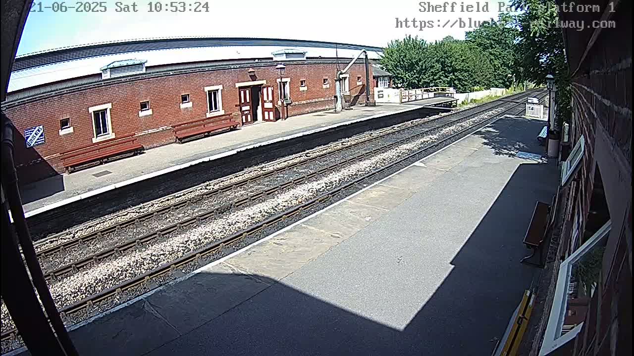 A sunny view of a train station platform with a brick building, benches, and train tracks.