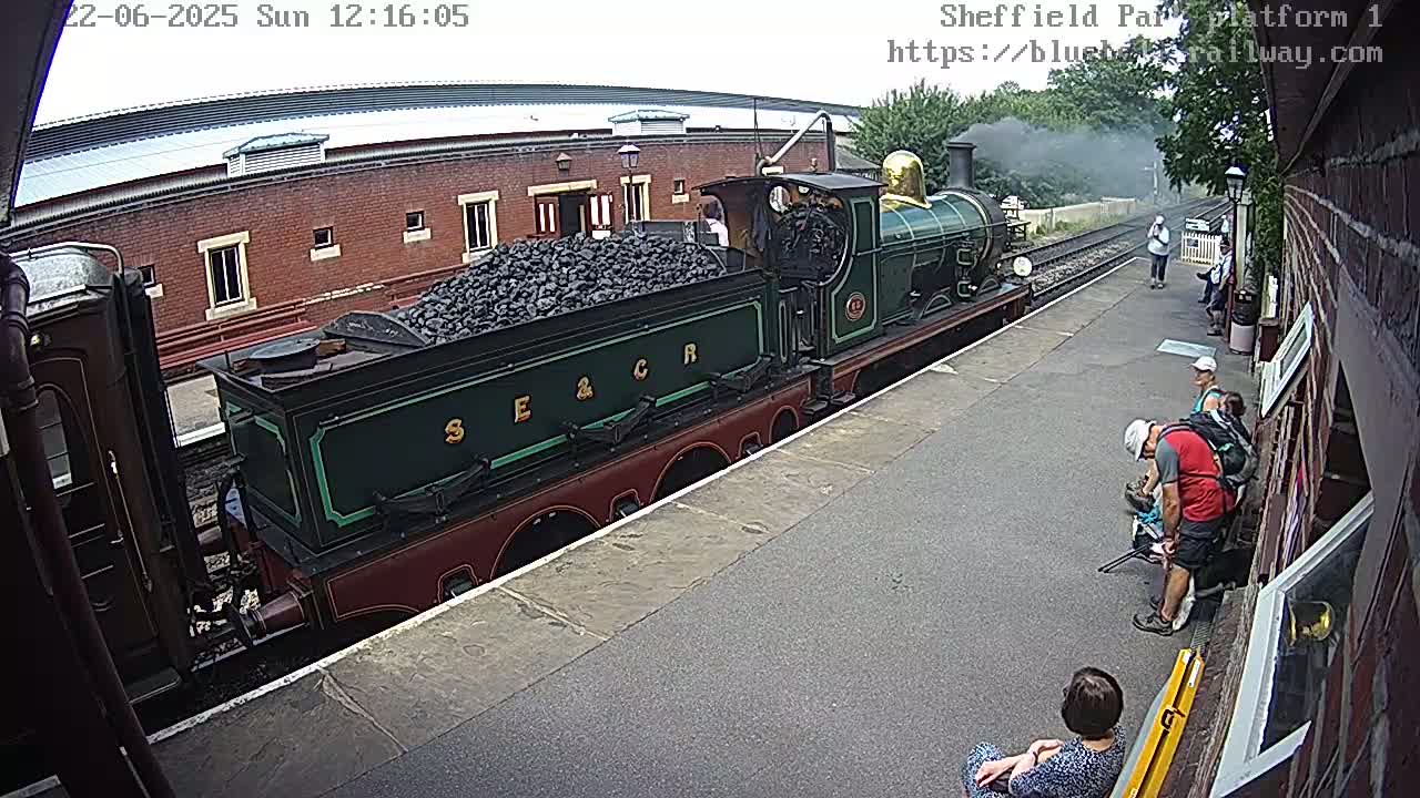 A green and maroon steam train loaded with coal sits at a station platform on a partly cloudy day.