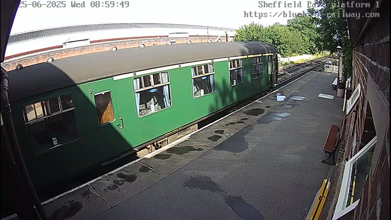 A dark green train car sits at a station platform on a partly sunny day with some wet spots on the ground.