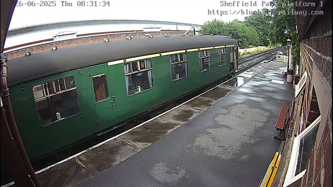 A dark-green railway carriage sits at a station platform on a damp, overcast day.