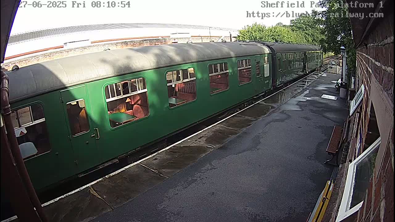 A long, dark-green passenger train sits at a wet train platform on a partly cloudy day.