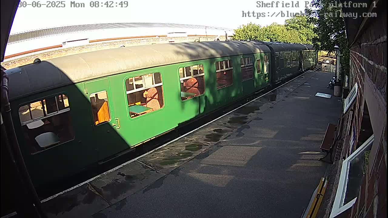 A long green train is parked at a station platform on a partly sunny day with wet pavement.