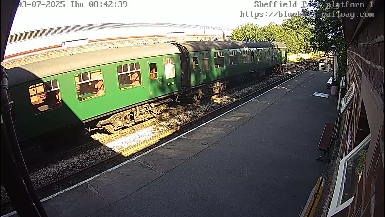 A long green passenger train sits on a railway track in bright sunlight.