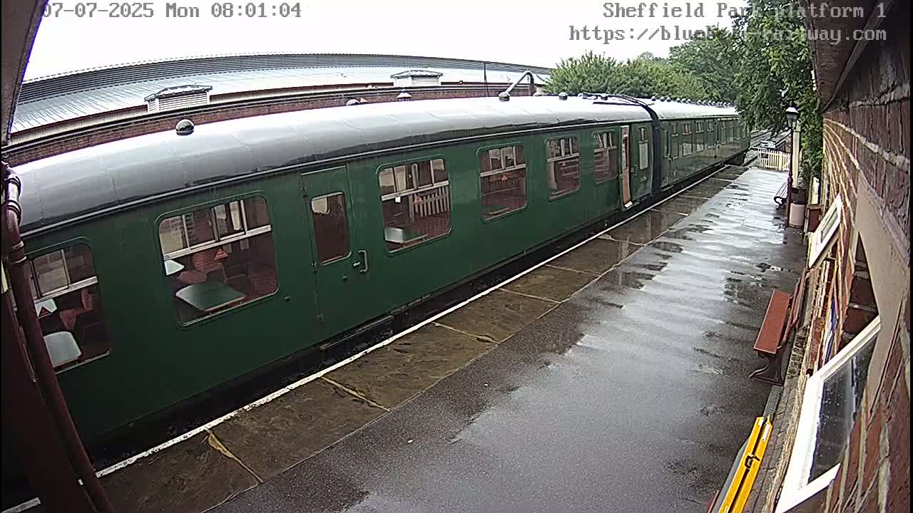 A long, dark-green passenger train sits at a rain-slicked platform next to a brick building.