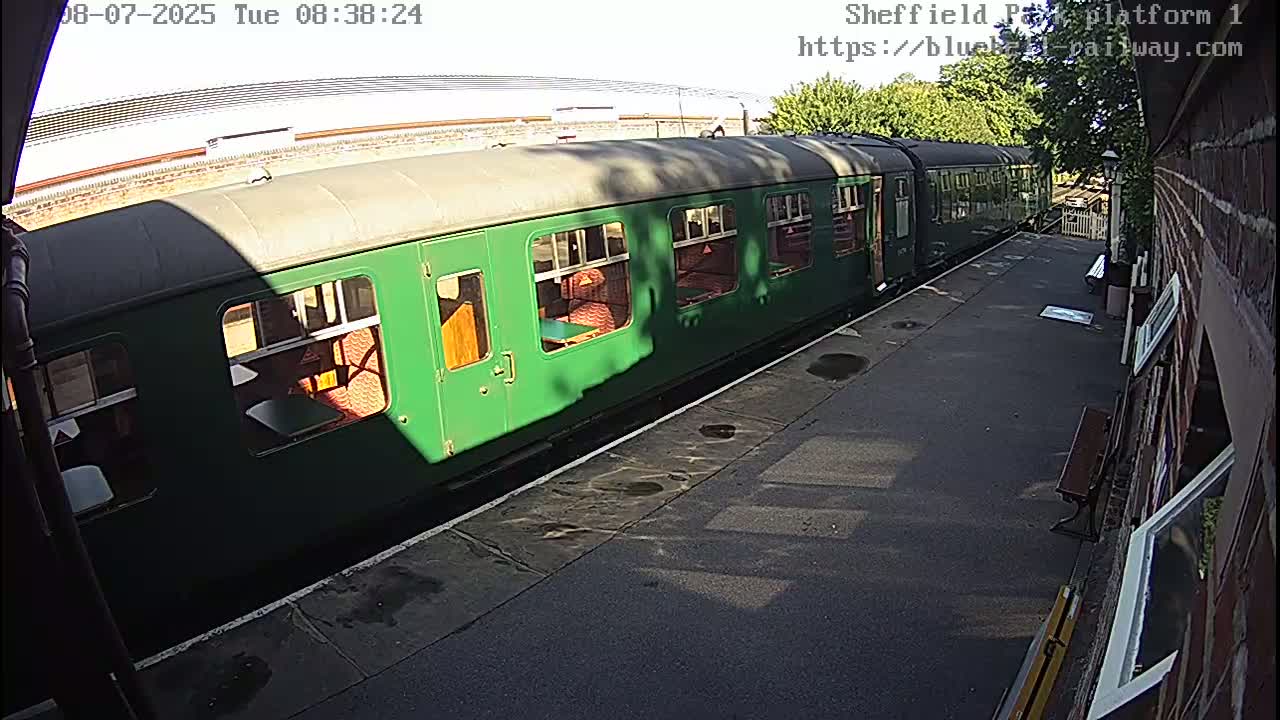 A long, dark-green passenger train sits at a station platform on a sunny day.