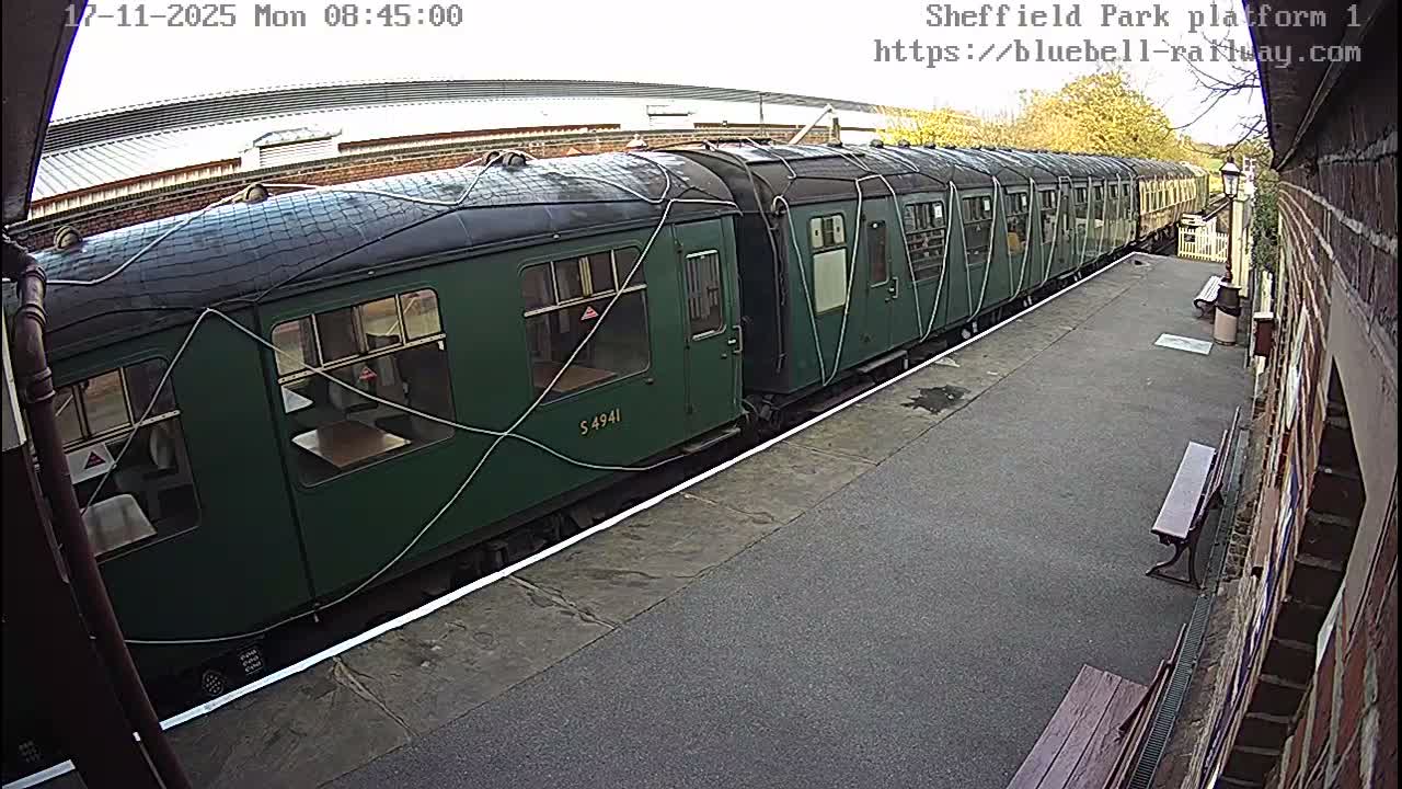 A long, dark green train with white cables draped over its top and sides rests at an outdoor station platform on a bright, clear day.