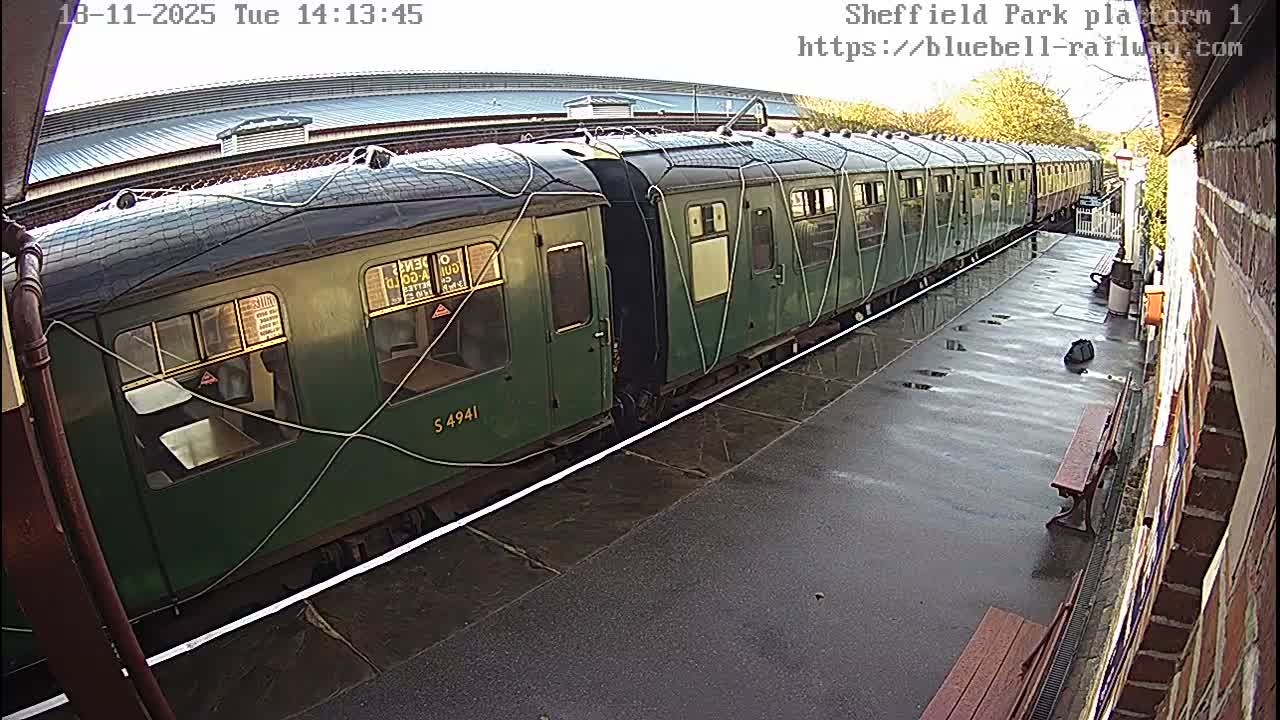 A long, dark green train with white cables draped over its top and sides rests at an outdoor station platform on a bright, clear day.