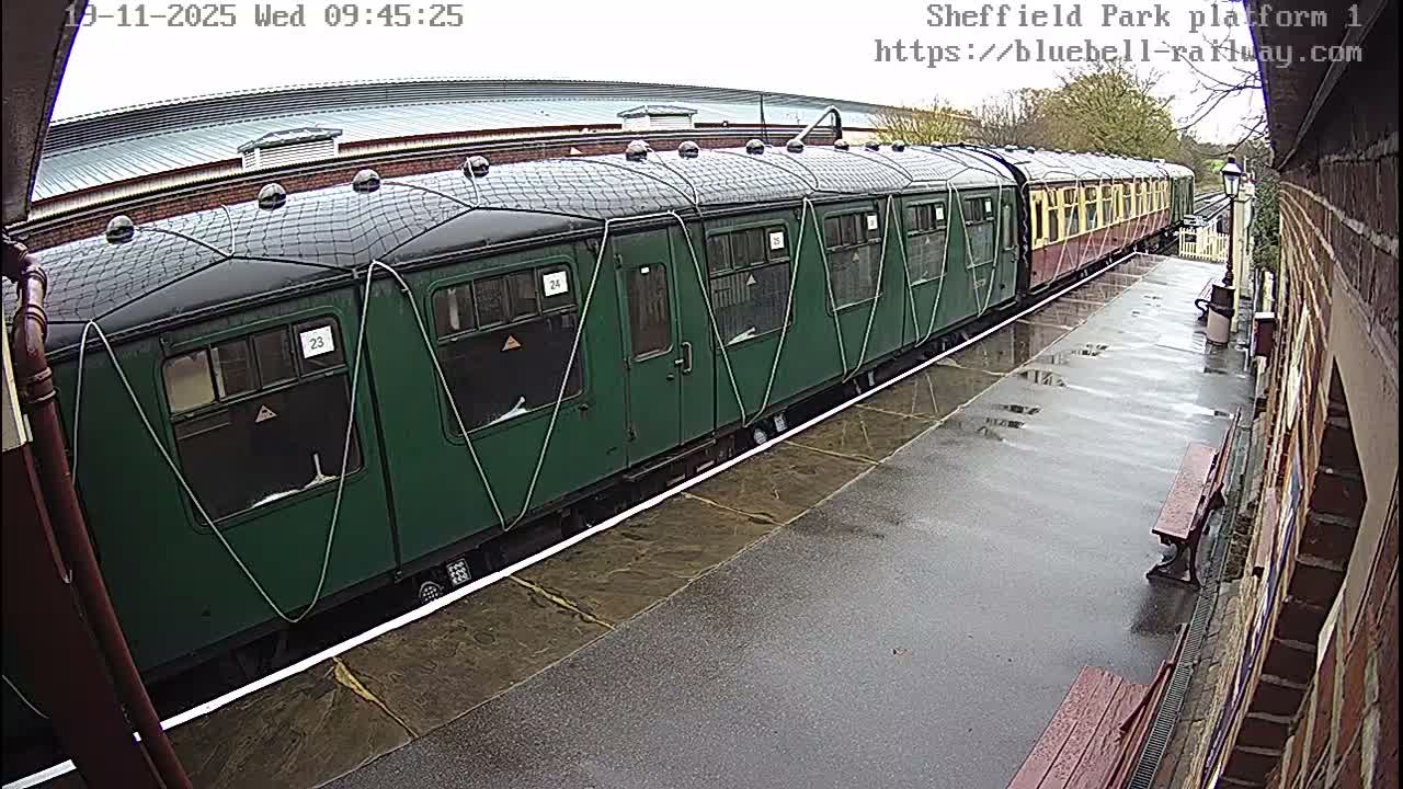 A long green and yellow train sits alongside a wet, empty platform at an outdoor station on an overcast day.