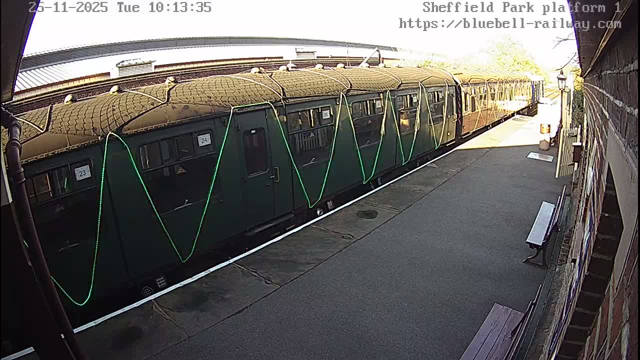 A dark green train, adorned with zig-zag green lights, sits at an outdoor station platform under bright sunny skies, with a brick building and a bench visible to the right.