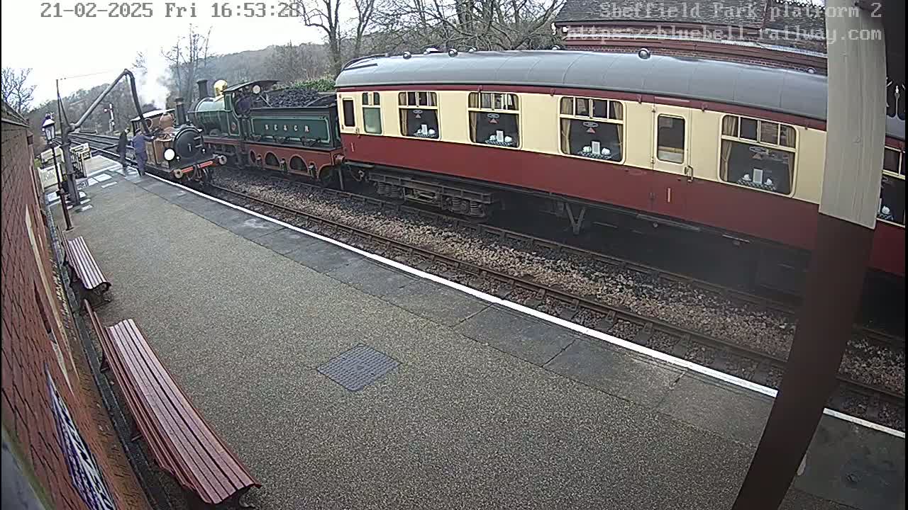 A steam train coupled to a passenger carriage sits at a station platform on an overcast day.