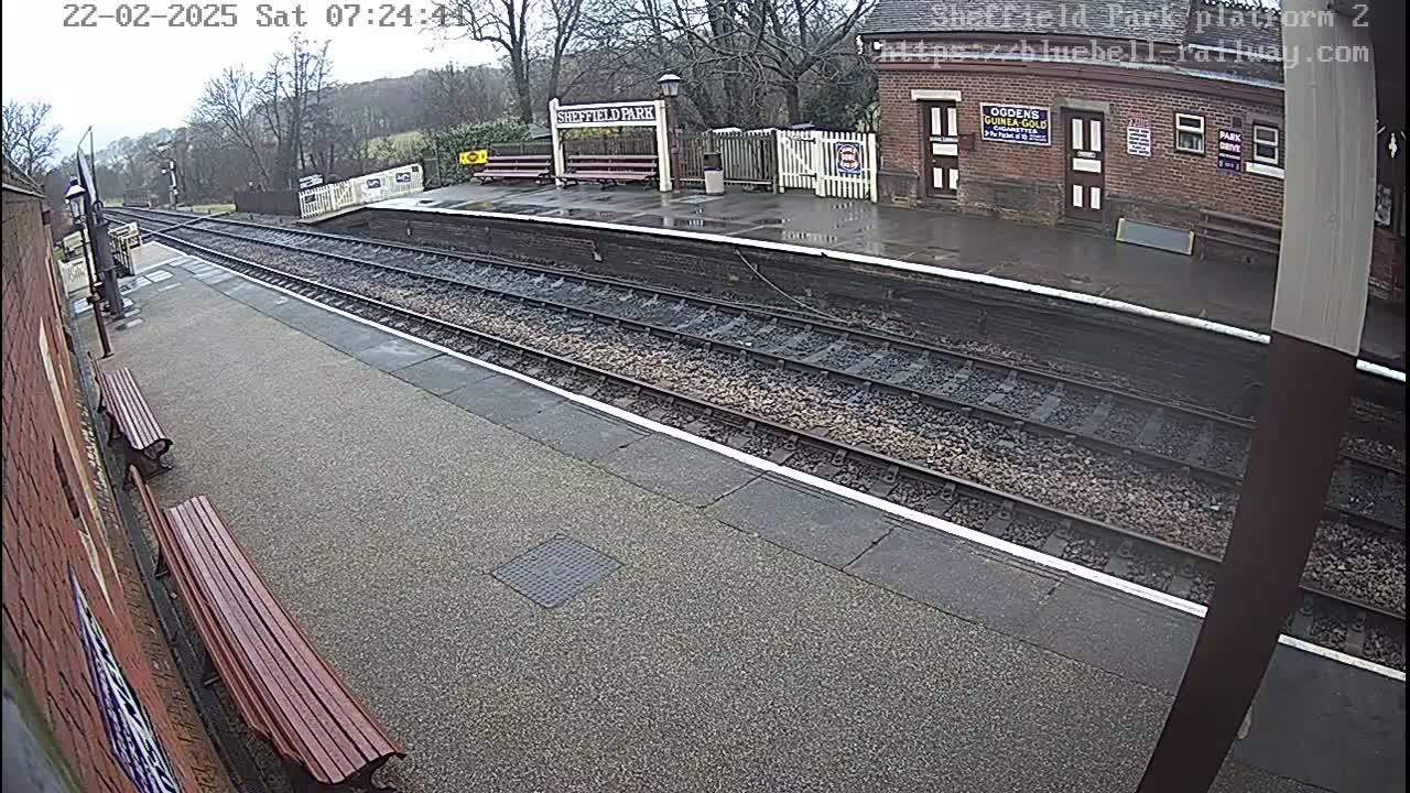 A wet train platform with two wooden benches and train tracks extending into the distance, under an overcast sky.