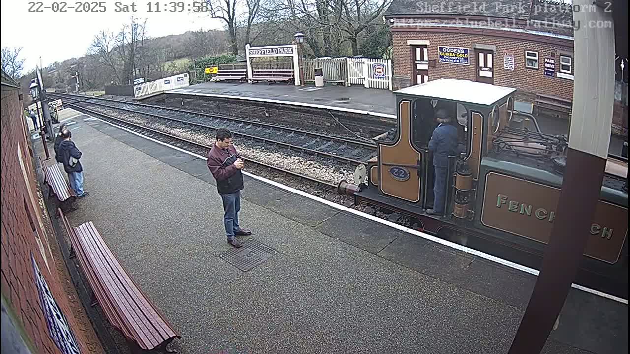 A small train is stopped at a railway station platform on an overcast day, with several people standing nearby.
