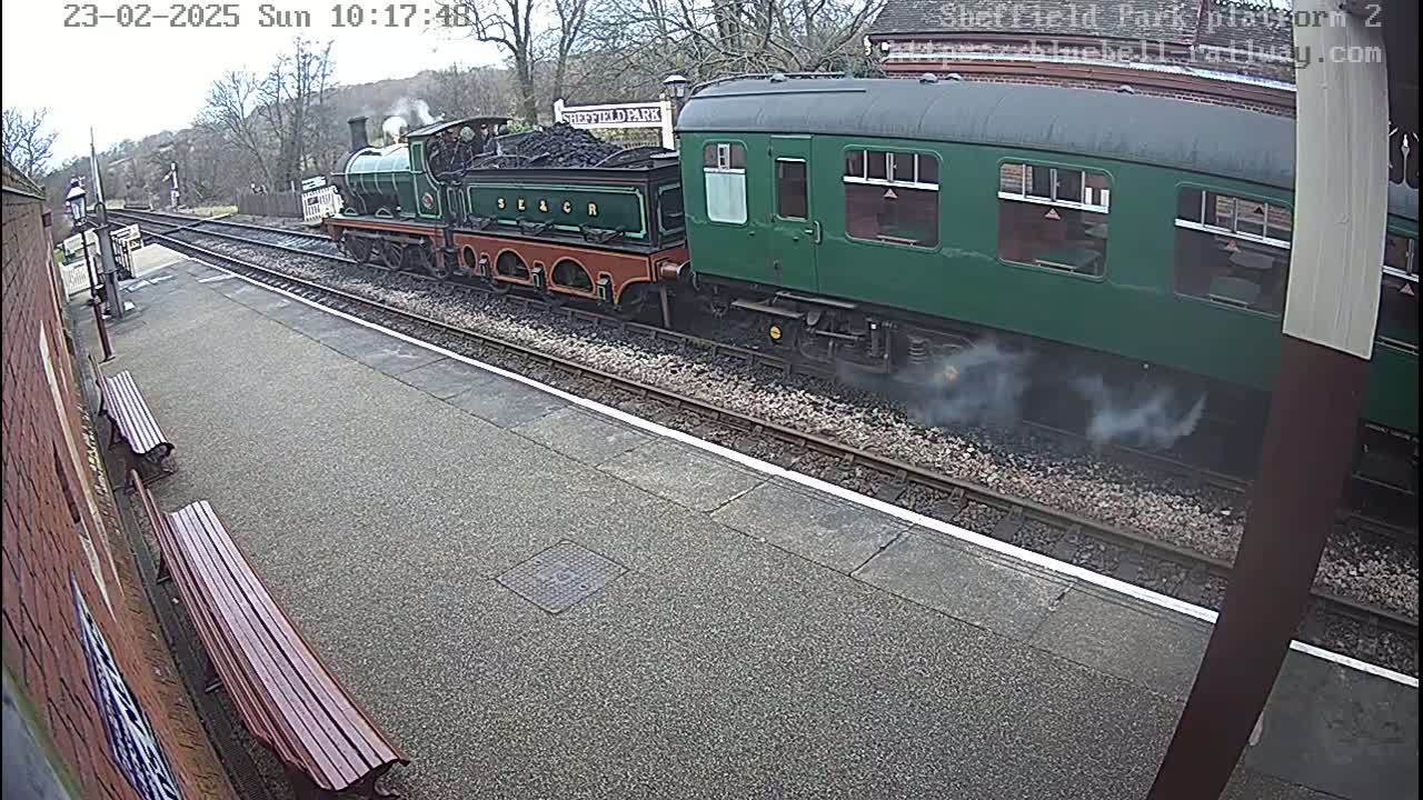 A steam train and a passenger car pull into a train station on an overcast day.