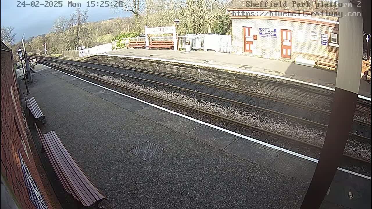 A sunny view of a train platform with two benches, railroad tracks, and a brick building in the background.