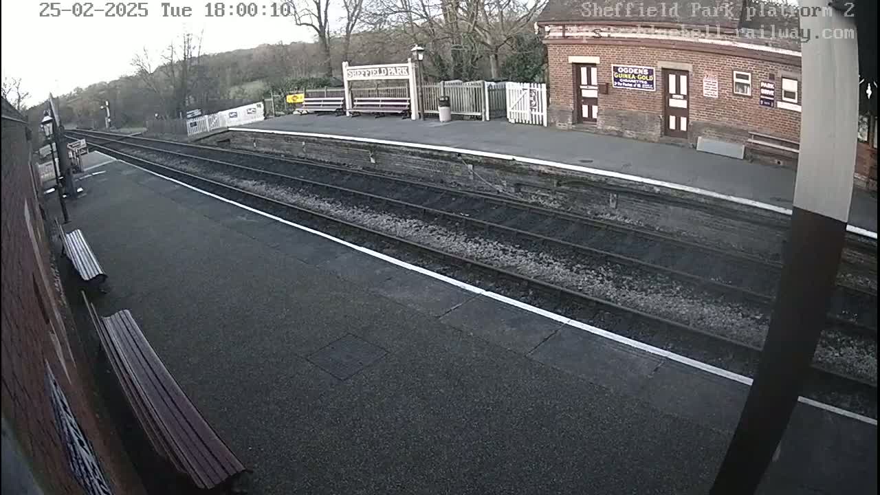 A mostly empty train platform with train tracks and a brick building is visible on an overcast day.