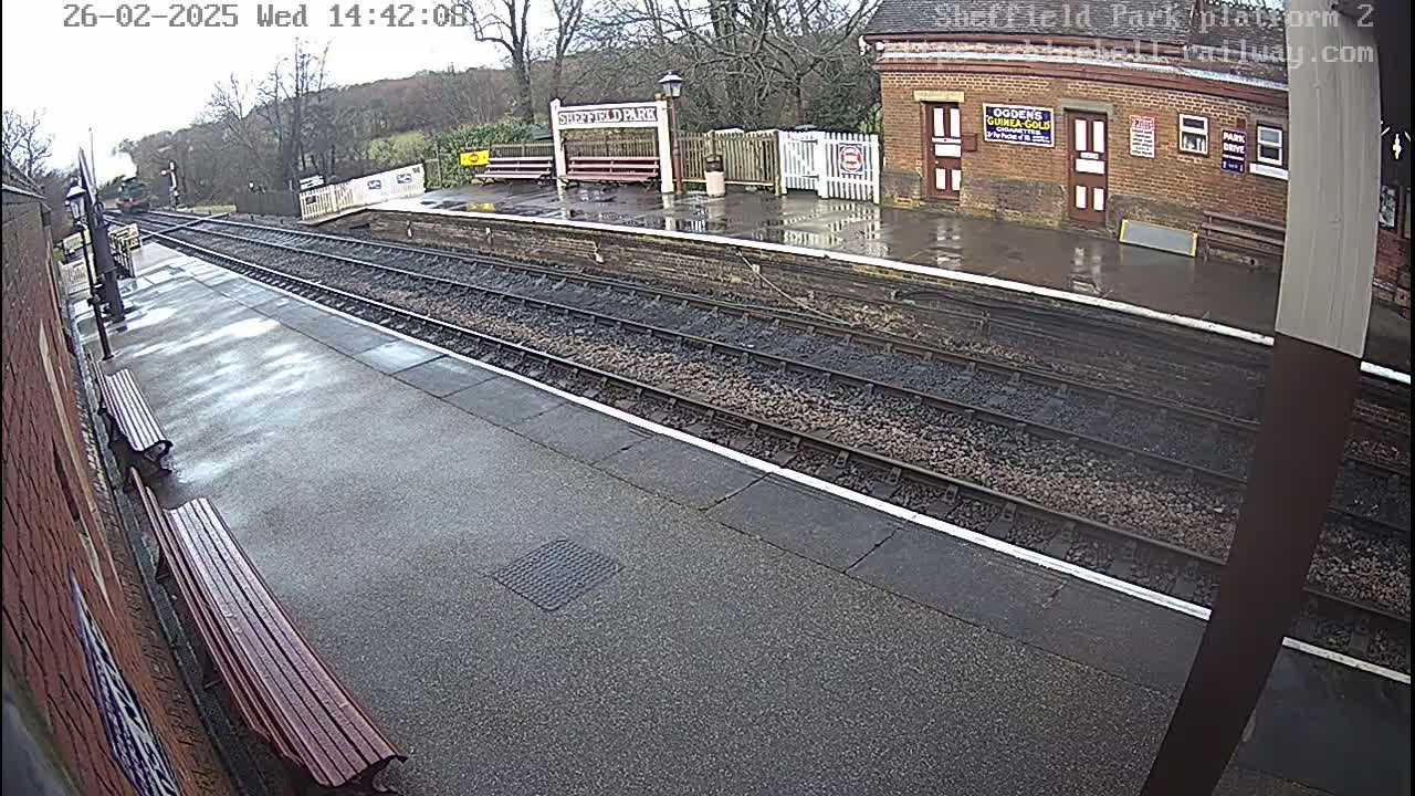 A wet train platform with two benches and train tracks extending into the distance under an overcast sky.