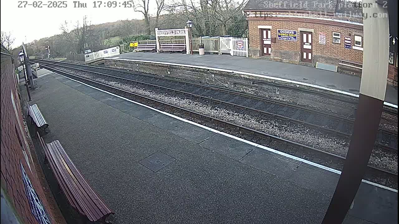 A mostly empty train platform on an overcast day, with two sets of train tracks and a brick building visible in the background.