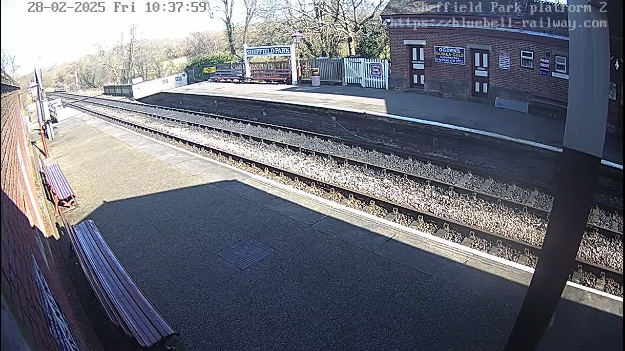 A sunny view of a train station platform with two benches, railway tracks, and a building in the background.