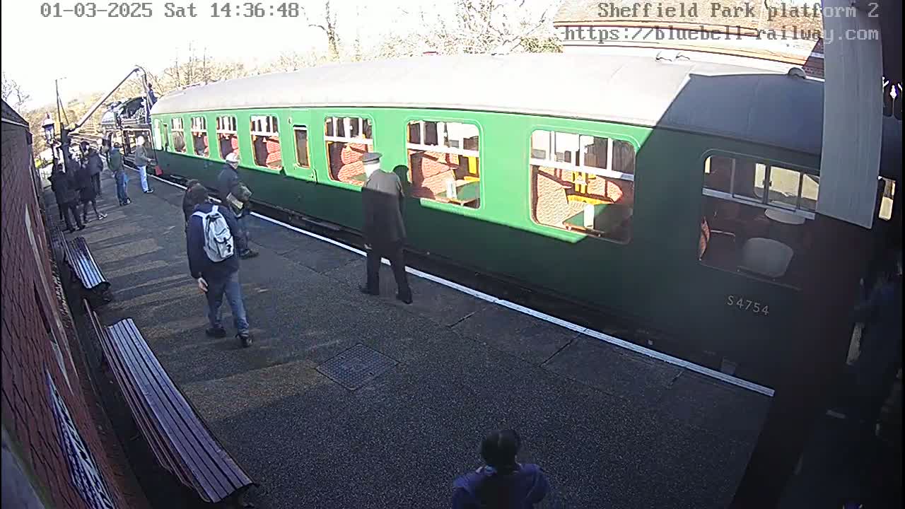 A green passenger train car is at a train station platform on a sunny day, with several people standing nearby.