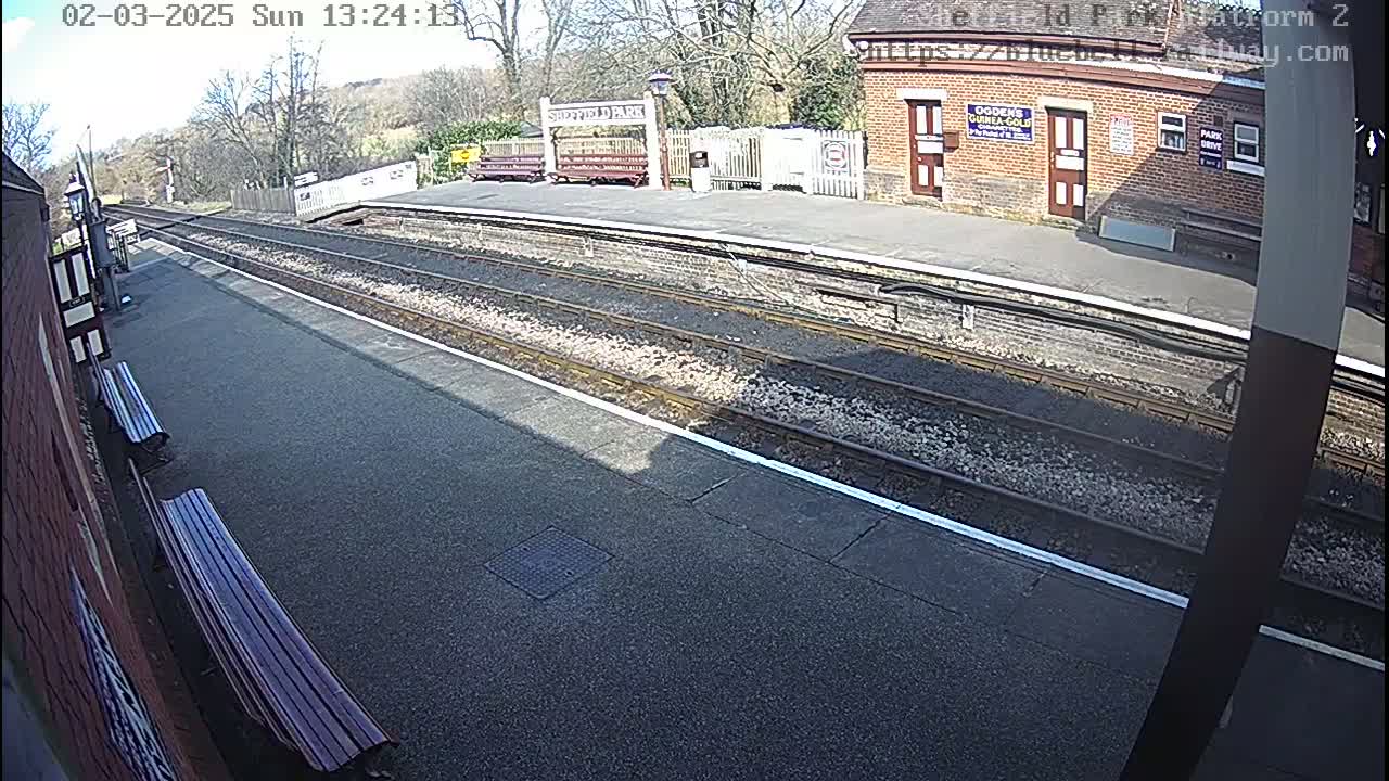 A sunny view of a train station platform with two benches, multiple train tracks, and a brick building.