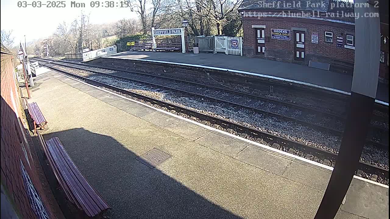 A sunny view of a train platform with two parallel sets of tracks, benches, and a brick station building.