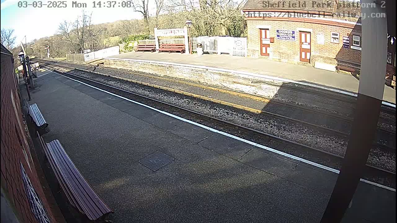 A sunny view of a train platform with two parallel train tracks, benches, and a brick building.