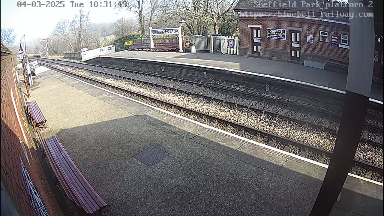 A mostly sunny view of a deserted train platform with two benches and a brick building in the background.
