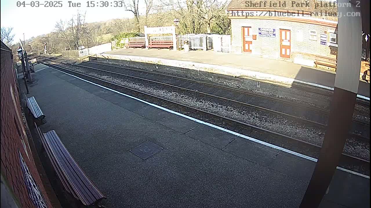 A sunny view of a mostly empty train platform with two benches, train tracks, and a brick building.