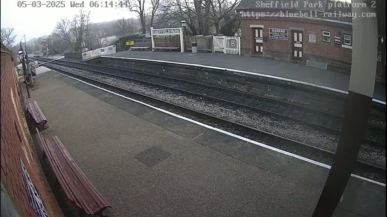 A train platform with empty tracks on an overcast day, two benches, and a brick building visible in the background.