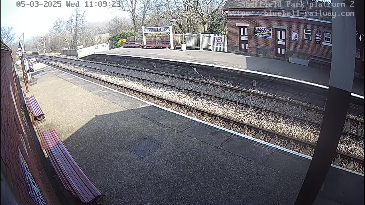 A train platform on a sunny day with two benches, train tracks, and a brick building.